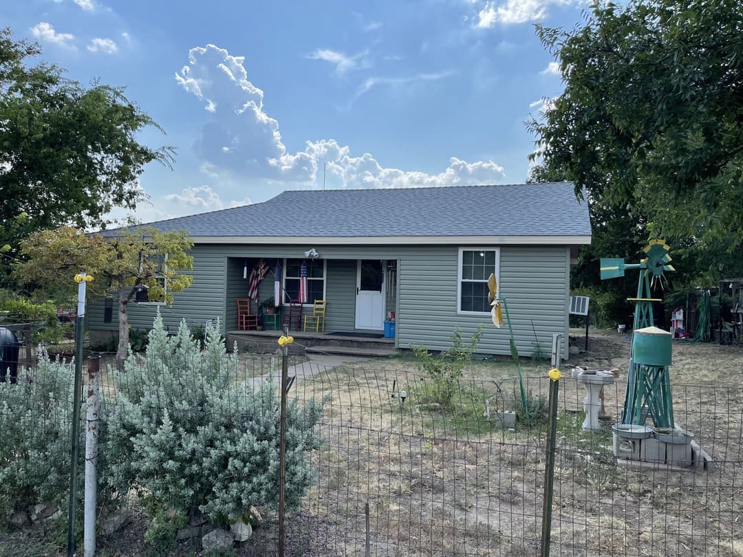 Single-story house with a porch, surrounded by greenery and blue sky with clouds.
